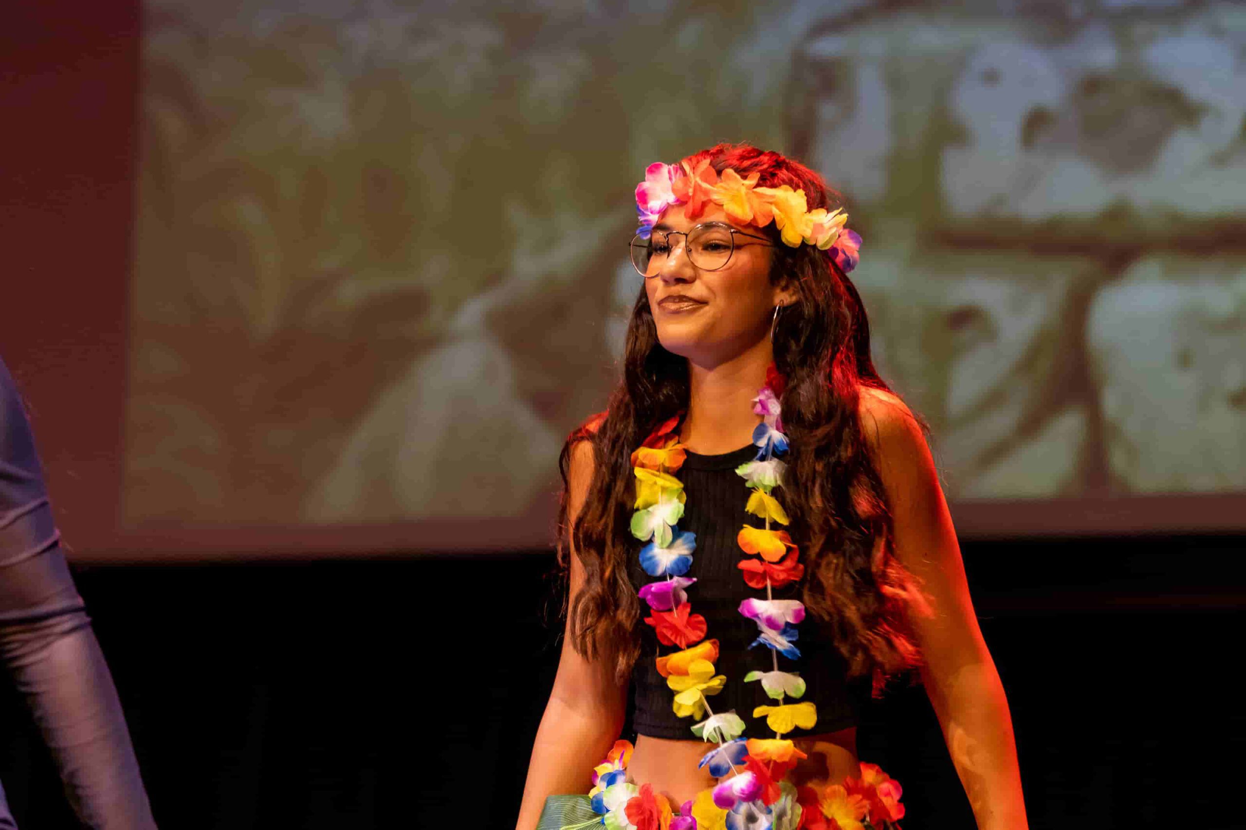 Bailarina de musical con corona y collar de flores coloridas en el escenario.