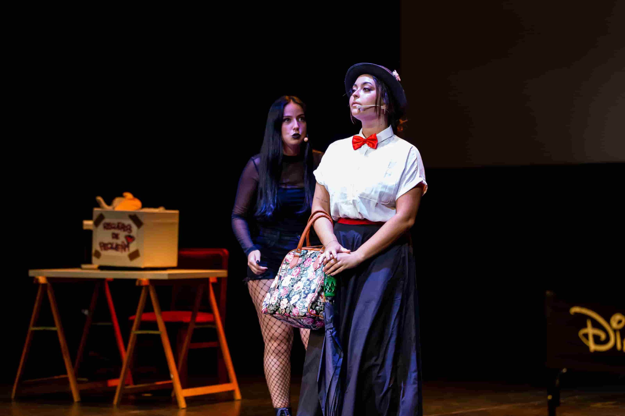 Dos actrices en una escena teatral, una lleva un bolso estampado y ropa de época.